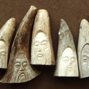 May include: Close-up of several carved antler pendants. Each pendant features a detailed face with a long beard and closed eyes. The pendants are various shades of cream and brown, with a hole at the top for stringing.