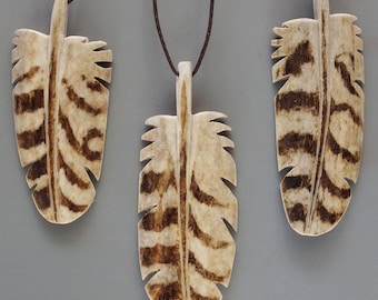 Op de afbeelding: Drie veervormige hangers met bruine en crèmekleurige patronen hangen aan bruine koorden. De hangers hebben een natuurlijke, organische uitstraling, met een getextureerd oppervlak en een gedetailleerd veermotief. De achtergrond is effen grijs.