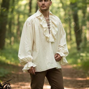 May include: Cream-colored long-sleeve shirt with ruffled collar and cuffs, and a laced neckline. The shirt is paired with brown pants and boots. The image is set in a forest, with the "MEDIECRAFT" logo in the bottom left corner.