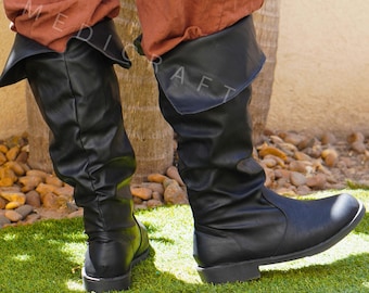 May include: Black leather knee-high boots with a folded-over top. The boots are worn with rust-colored pants and are on a green grassy surface. The boots have a slight heel and a rounded toe. The logo "MEDIECRAFT" is visible.