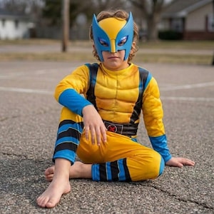 May include: A yellow and blue superhero costume featuring a moulded mask with pointed ears, a long-sleeved top with padded chest detailing, and matching trousers. The outfit includes black accents on the sleeves and legs, and a belt with an X-shaped emblem.