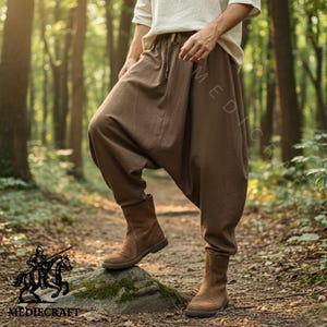 May include: Brown harem pants with a drawstring waist and matching brown boots. The pants have a loose, baggy fit. The boots are ankle-high and made of a suede-like material. The image is set in a forest.
