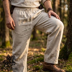 Könnte beinhalten: Beige Leinenhose mit Kordelzug in der Taille. Die Hose ist Teil eines passenden Sets, und das Model trägt ein langärmliges Hemd. Die Hose hat eine lockere Passform und ist für den Freizeitgebrauch geeignet. Das Logo "MEDIECRAFT" ist unten links sichtbar.