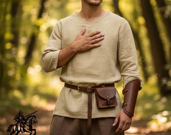 May include: A beige linen tunic with a V-neck, paired with brown pants and a leather belt. A small brown leather pouch hangs from the belt. A brown leather arm guard is worn on the right arm. The image includes the logo of Mediecraft.