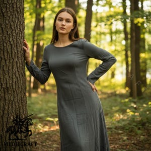 May include: A long-sleeved, grey dress with a round neckline. The dress has a fitted bodice and a flowing skirt. The model is standing in a forest setting. The logo "MEDIECRAFT" is visible in the bottom left corner.