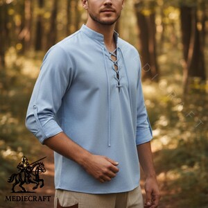May include: A pale blue tunic shirt featuring a stand-up collar and a laced front. The sleeves are three-quarter length with buttoned cuffs. The shirt is made of a soft material. The logo "MEDIECRAFT" is visible at the bottom left.