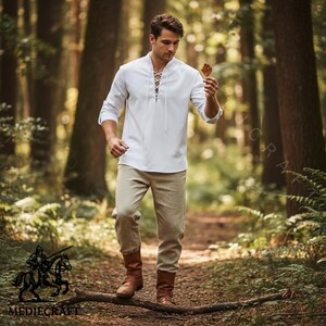 May include: A white long-sleeve shirt with a lace-up neckline, tan trousers, and brown boots. The shirt has rolled-up sleeves. The person is walking on a path in a forest. The image includes the logo "MEDIECRAFT".