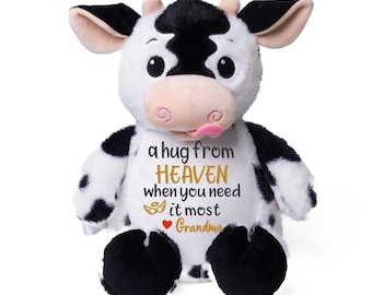 Op de afbeelding: Een pluche koe-speeltje met zwart-witte vacht, een roze snuit en een uitgestoken tong. Het witte borstgedeelte is geborduurd met de tekst 'a hug from HEAVEN when you need it most', gevolgd door een klein engelvleugel-icoontje en 'Grandma'.