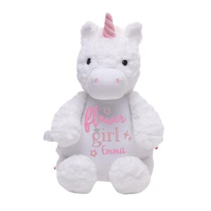 May include: A white plush unicorn with a pink horn and accents. The unicorn has the words "flower girl" and the name "Emma" embroidered on its chest in pink. The unicorn is a soft, cuddly toy.