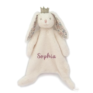 May include: A cream-colored plush rabbit security blanket featuring long ears with a floral pattern on the inner side, a small gold crown on its head, and the name Sophia embroidered in a dusty rose script on the center of the body.