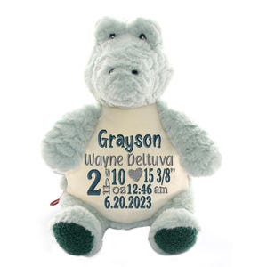 May include: A light blue plush hippopotamus with a cream-colored belly featuring embroidered text: Grayson, Wayne Deltuva, 2 lbs 10 oz, a grey heart, 15 3/8", 12:46 am, and 6.20.2023. The stuffed animal has dark green textured fabric on its feet.