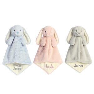 May include: Three plush security blankets featuring soft bunny heads with long ears. The blankets are light blue, pale pink, and light gray with cream-colored undersides. Each blanket is embroidered with a name: Brody James, Jodi, and John.
