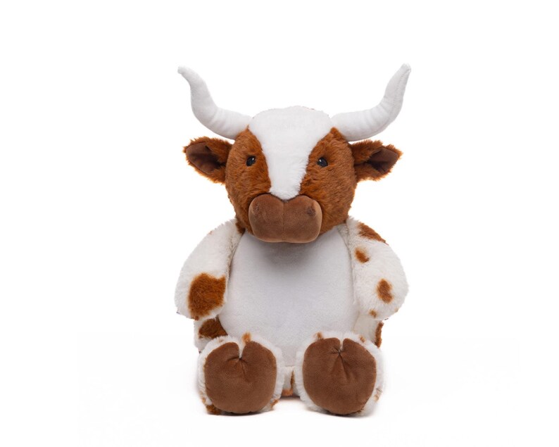 A stuffed toy cow with white horns and brown spots sits against a white background.