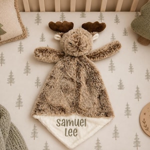 May include: A plush moose security blanket with soft brown faux fur, dark brown antlers, and a cream-colored fabric corner embroidered with the name Samuel Lee in dark gray thread, centered on a light-colored fabric with a green tree pattern.