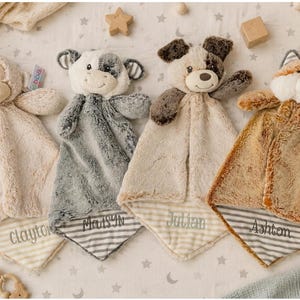 May include: Four plush security blankets featuring animal heads: a monkey, a cow, a dog, and a fox. Each soft, fuzzy blanket has a striped fabric corner embroidered with the names Clayton, Madsyn, Julian, and Ashton in gray thread.