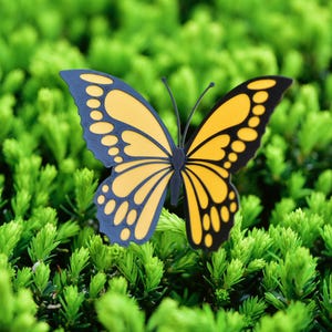 May include: A decorative butterfly with yellow and black wings, and blue wing tips, rests on a bed of green foliage. The butterfly's wings have a detailed pattern. The image is a close-up shot, highlighting the butterfly's intricate design and the vibrant green background.