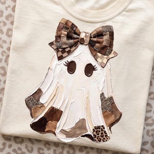 Può includere: Una t-shirt color crema con una grafica centrale raffigurante un fantasma che indossa un grande fiocco a quadri marrone. Il design del fantasma incorpora varie texture marroni e beige, inclusi motivi leopardati e velluto a coste.