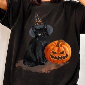 Może przedstawiać: Czarny t-shirt z krótkim rękawem z nadrukiem graficznym przedstawiającym czarnego kota w spiczastym kapeluszu czarownicy ozdobionym pomarańczowymi gwiazdami i księżycami, obok pomarańczowej dyni z wyciętą, ząbkowaną, uśmiechniętą twarzą.