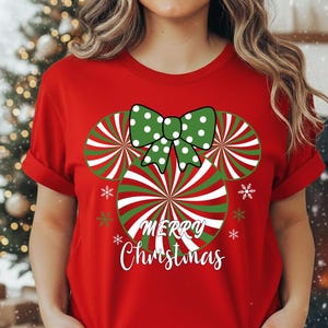 May include: A red t-shirt featuring a Christmas-themed design. The design includes a bow and Mickey Mouse-shaped candy canes with the words "Merry Christmas" in white. The shirt is short-sleeved.