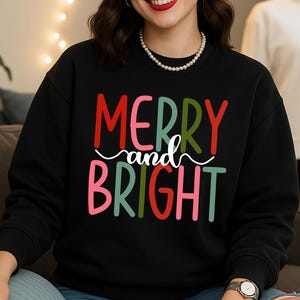 Könnte beinhalten: Schwarzes Sweatshirt mit den Worten "MERRY and BRIGHT" in roter, grüner, rosa und weißer Schrift. Die Person trägt eine Perlenkette und eine silberne Uhr.