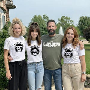 May include: Four people pose for a photo. Three wear white t-shirts with a black graphic of The Child from The Mandalorian. The fourth person wears a green t-shirt with "The Dadalorian" and "This is the Way" printed on it.