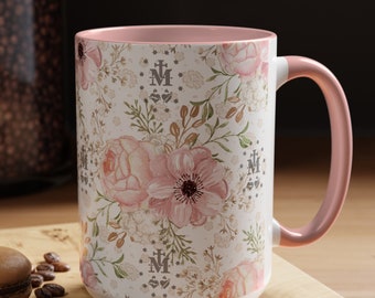 May include: A white ceramic mug with a pink rim and handle. The mug features a floral pattern with pink roses and white flowers. The pattern also includes a repeating symbol of a stylized "M" with a cross and heart.