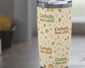 May include: A stainless steel travel tumbler with a cream-colored background and a repeating pattern of religious symbols and rainbows. The tumbler has the text "Catholic, but chill" in a brown, stylized font.