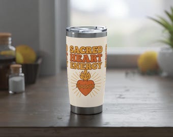 May include: A cream-coloured travel tumbler featuring the text "Sacred Heart Energy" in orange and brown. The design includes an orange heart with a flame and sunburst rays. The tumbler has a silver lid and sits on a wooden surface.