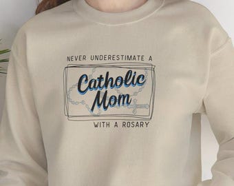 May include: Beige sweatshirt with the text "NEVER UNDERESTIMATE A Catholic Mom WITH A ROSARY" in black and blue. The text is inside a rectangular frame with a rosary design.