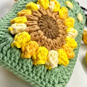 May include: A handmade crochet pouch in a light green color, featuring a large sunflower design. The sunflower has yellow, cream, and brown petals and a brown center. The pouch has a textured appearance, with a yellow tassel attached.