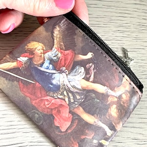 May include: A small, square coin purse featuring a Renaissance-style painting of a figure with wings, holding a sword. The artwork depicts a battle scene with figures in red, blue, and gold. The purse has a black zipper and a silver charm.
