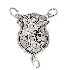 May include: A silver pendant depicting Saint Michael the Archangel. The shield-shaped pendant has key rings at the top and bottom. The image shows Saint Michael with wings, a halo, and a sword, standing over a defeated figure. The pendant is made of a silver-toned metal.