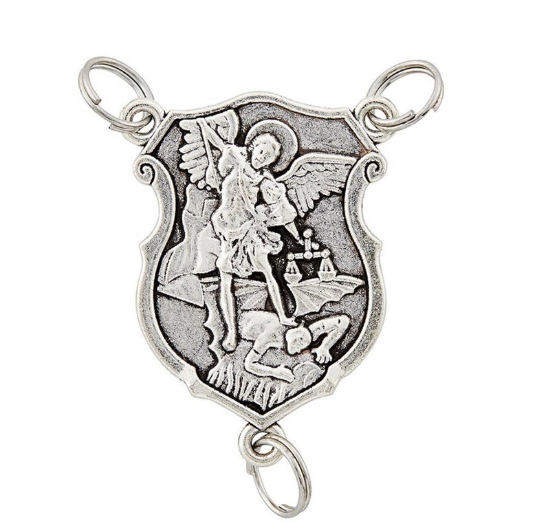 May include: A silver pendant depicting Saint Michael the Archangel. The shield-shaped pendant has key rings at the top and bottom. The image shows Saint Michael with wings, a halo, and a sword, standing over a defeated figure. The pendant is made of a silver-toned metal.