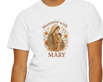 May include: White t-shirt featuring a graphic of a woman holding a child, surrounded by floral elements. The text "Mommin' with MARY" is arched above and below the image. The design is in shades of brown, green, and orange.