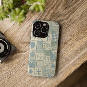 Peut inclure: Une coque de téléphone bleu clair et crème avec un motif patchwork représentant des symboles religieux tels que des croix, des poissons et des cœurs. La coque est posée sur une surface en bois, avec un appareil photo et une plante en arrière-plan.