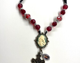 Op de afbeelding: Een rood en zwart rozenkrans met een cameo van de Maagd Maria en een kruisbeeld hanger. De rozenkrans kralen zijn gemaakt van glas en de hanger is gemaakt van metaal.