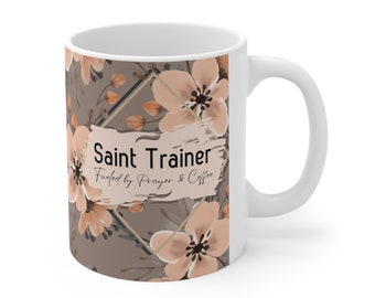 Könnte beinhalten: Weiße Keramik-Tasse mit floralem Design in Pfirsich- und Brauntönen. Der Becher zeigt den Text "Saint Trainer Fueled by Prayer & Coffee". Ideal für Kaffee oder Tee.