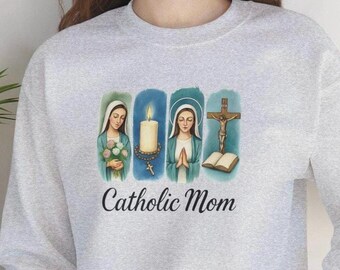 May include: A light gray sweatshirt featuring a graphic with religious imagery. The graphic includes illustrations of a woman with flowers, a candle, a woman praying, and a crucifix. The words "Catholic Mom" are printed below the images.