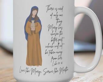 Könnte beinhalten: Weißer Keramikbecher mit einer Illustration einer Frau in blauem Kopftuch und goldenem Gewand, die eine Vase hält. Der Becher enthält den Text "Es bedarf nur eines..." und "Liebe wie Maria, diene wie Martha."