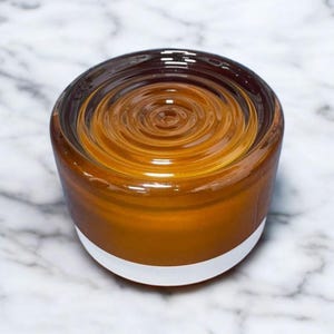 Pode incluir: Um frasco de vidro redondo âmbar com uma base branca. O conteúdo do frasco tem um design em espiral. O frasco está sobre uma superfície de mármore branco e cinza. O frasco é provavelmente uma vela ou um recipiente para um produto cosmético.