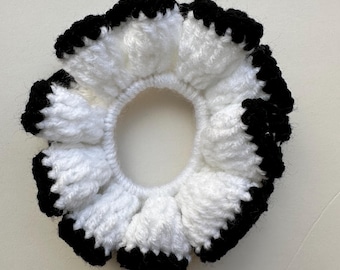 May include: A handmade scrunchie with a flower-like design. The scrunchie is crocheted with white yarn and has a black border. The hair accessory is designed to hold hair in a ponytail or bun.