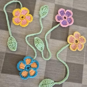 May include: Crocheted flower bookmarks in various colors, including yellow, pink, blue, and purple. Each flower has a green stem and leaves. The handmade bookmarks are arranged on a checkered surface.