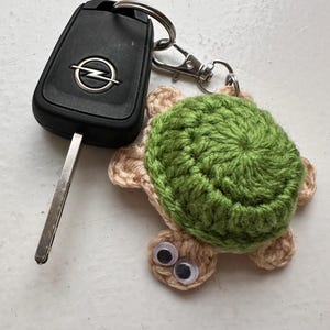 May include: A handmade crochet turtle keychain with a green shell and tan body, attached to a silver keyring. The keychain is next to a black car key with the Opel logo.