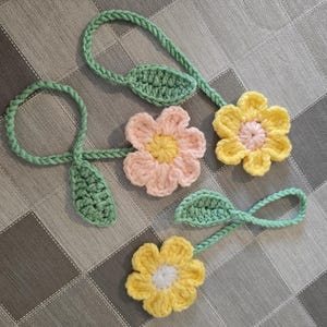 以下が含まれることがあります： 黄色、ピンク、緑色の手編みの花と葉の飾り。花は花びらとは異なる中心の色をしています。葉と茎は緑色です。グレーと白のチェック柄の表面に配置されています。