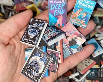 Puede incluir: Una colección de libros en miniatura con varios títulos y diseños de portada. Algunos títulos visibles incluyen "Deep End", "Dating Dragons", "Nightbane" y "Order of Swans". Los libros presentan una variedad de colores y estilos, sugiriendo un tema de fantasía o romántico.