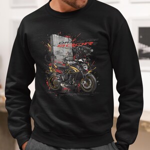 Pode incluir: Uma camisola preta de manga comprida com uma impressão gráfica de uma mota e o texto "DRAGSTER 800RR" em vermelho e branco. O design inclui um fundo de parede de betão com salpicos de tinta vermelha e amarela e uma silhueta de pantera.