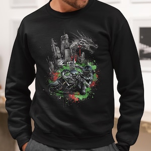 Op de afbeelding: Zwart sweatshirt met een grafisch ontwerp van een draak, een skyline en motorrijders. Het ontwerp bevat rode en groene verfspatten. Het sweatshirt heeft een ronde hals.