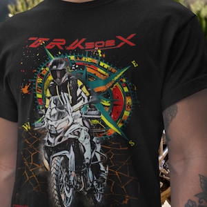 May include: A black t-shirt featuring a graphic print of a motorcyclist in a helmet and gear riding a white motorcycle. Behind the rider is a colorful compass design with the text TRKspeX in red at the top and orange cracked ground patterns.