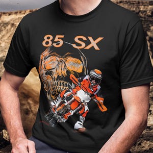 Puede incluir: Camiseta negra con un gráfico de calavera naranja y el texto "85 SX". La camiseta también muestra a un piloto de motocross con equipo naranja y blanco en una moto de cross.