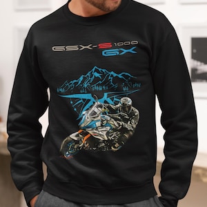 Pode incluir: Camisola preta com um gráfico de motocicleta e o texto "GSX-S 1000 GX". O design inclui um motociclista numa moto com montanhas no fundo. A camisola é de gola redonda.